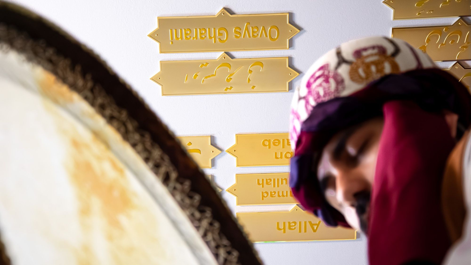 In the foreground, a man with his eyes closed is playing a frame drum; in the background, there are golden plaques on a wall.