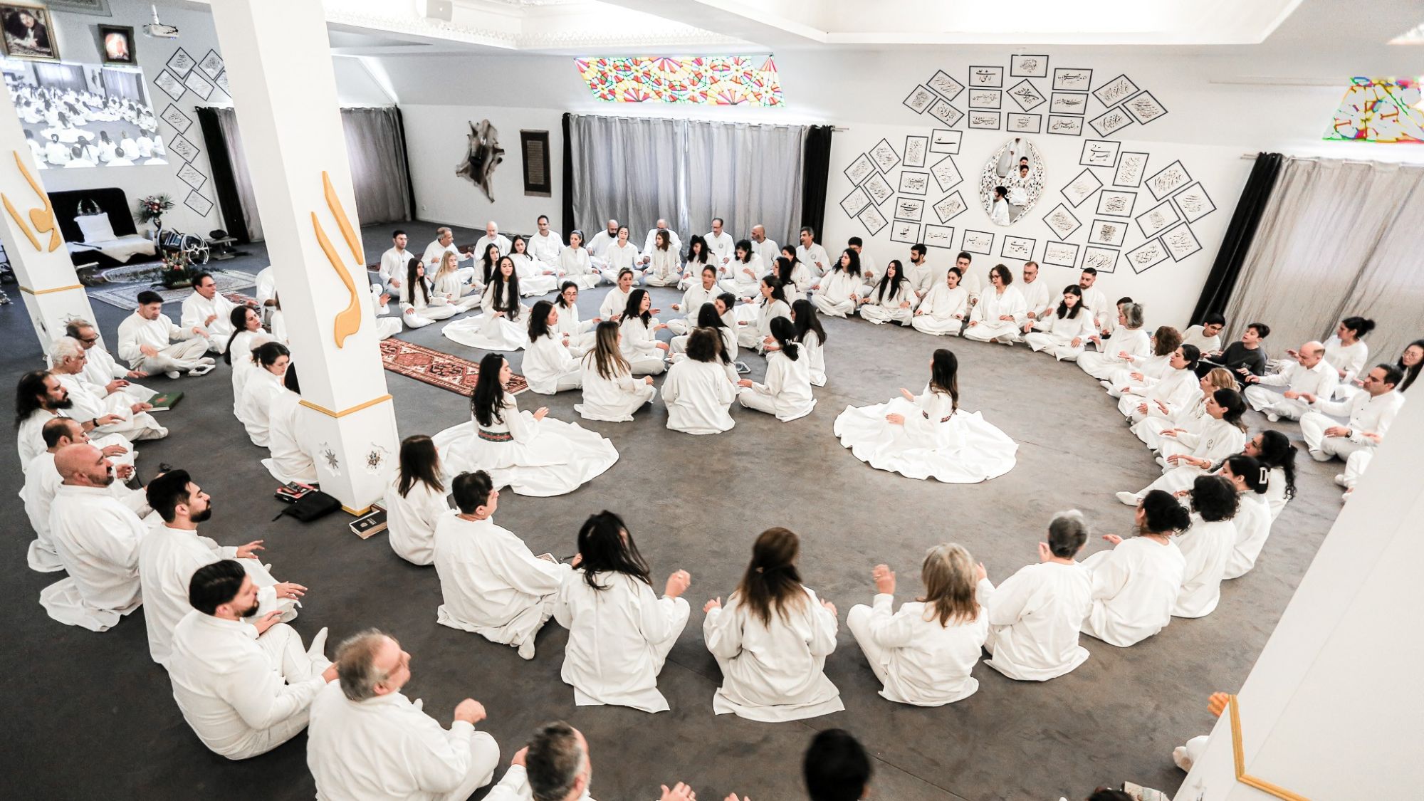 In large circles, people dressed in white are seated during a ceremony in a brightly lit room.