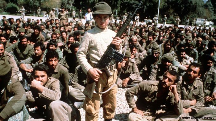 جندي إيراني صغير السن يرفع بندقية جي سي إيرانية الصنع أمام الكاميرا ويحيط به مئات الأسرى العراقيين من سنوات الحرب.