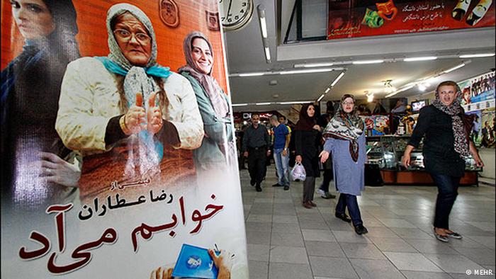 People shopping in a mall in Iran in the evening, a film poster in the foreground. Photo: MEHR