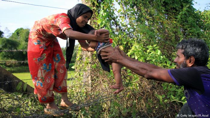 A Rohingya man passes an infant through a barbed wire border fence