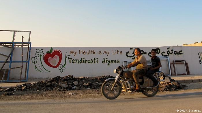 A man and a boy on a motorcycle ride past a mural in Kobani (photo: DW/Zurutuza)
