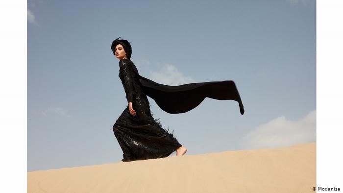 A woman in a black dress with a billowing scarf walks across a sand dune (photo: Modanisa)