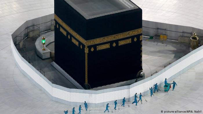 The imposing black square building of the Kaaba sits in the centre of the Grand Mosque in Mecca while a line of seemingly tiny men in green sanitation uniforms clean the usually busy floor of white tiles