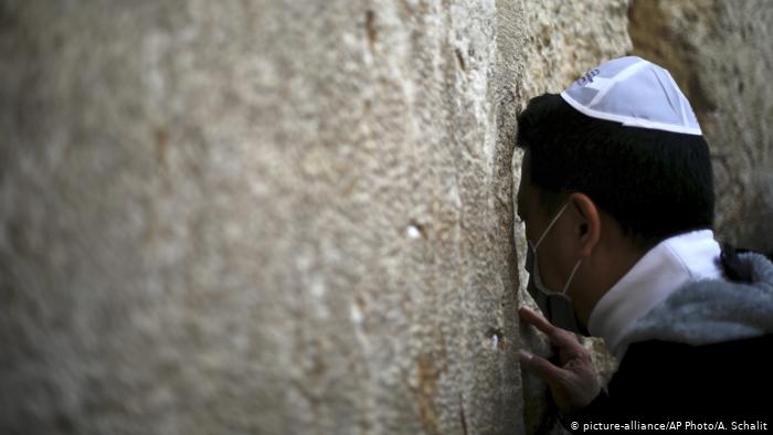 A man wearing a kippah and medical mask leans in to a crack in a large stone wall