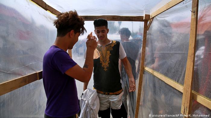 Inside a makeshift wooden frame lined with plastic, a young man sprays down another with a small spray bottle