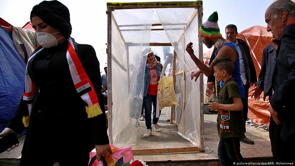 Demonstranten auf dem Tahrir-Platz in Bagdad gehen durch eine behelfsmäßige Desinfektionskabine, welche von der Protestbewegung zum Schutz gegen die Verbreitung des neuartigen Coronavirus aufgebaut wurde; Foto: picture-alliance/dpa/AP/K. Mohammed