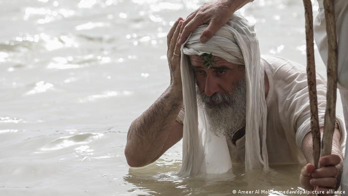 A person puts a hand on the head of an old man with a cloth on his head and holding onto a wooden stick, standing chest-deep in water