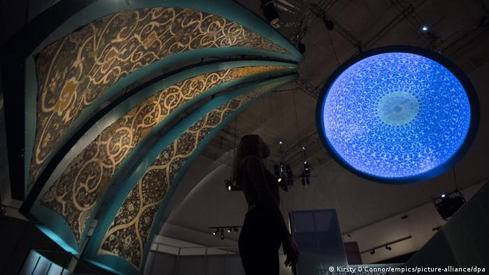 A museum staff member looks up at tilework and a bright blue dome projection in a darkened room at the 'Epic Iran' exhibition