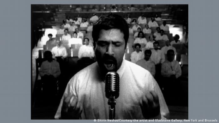 Black and white image of a man in front of a microphone with people in the background; an excerpt from Turbulent by Shirin Neshat as part of the Epic Iran exhibition