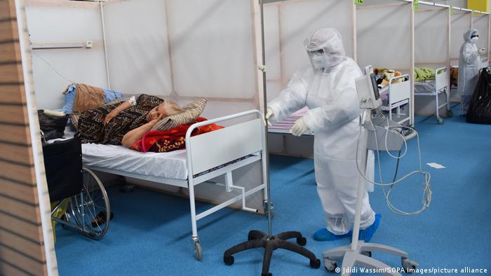 Hospital scene, masked person in white protective suit approaches a bed with a patient, more beds lined up in the background (photo: Jdidi Wassim/SOPA Images/picture-alliance)