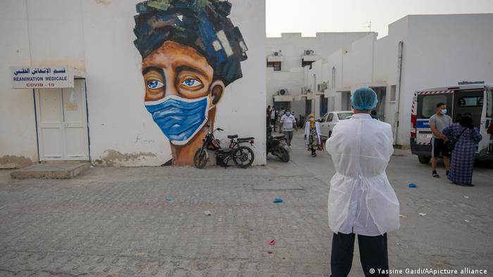 Person wearing a white lab coat and a green surgical cap watches some people walk among square white buildings, one with a large graffiti of a face with a mask (photo: Yassine Gaidi/AA/picture-alliance)