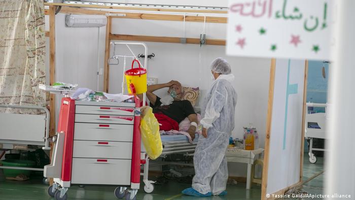 Person in a white protective suit looks at a patient in a bed in a makeshift cubicle, cart with medical appliances in the foreground (photo: photo: Yassine Gaidi/AA/picture-alliance)