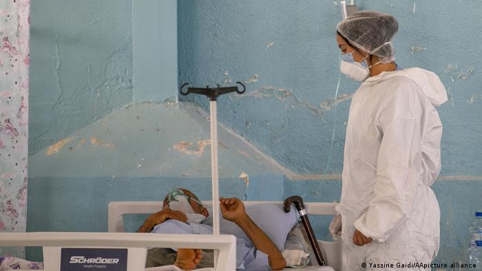 Person in a white protective suit, surgical cap and face mask looks at a patient in a bed in front of a rough pale blue wall (photo: Yassine Gaidi/AA/picture-alliance)