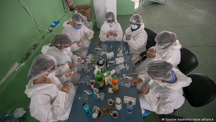 Seven people in protective suits, caps and masks sit around a table (photo: Yassine Gaidi/AA/picture-alliance)