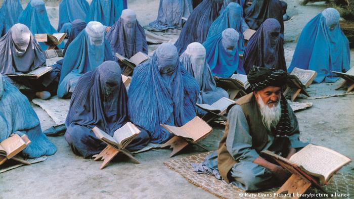 Rows of women wearing burkas and sitting on mats in the sand behind book rests, wih an elderly bearded man in the front row