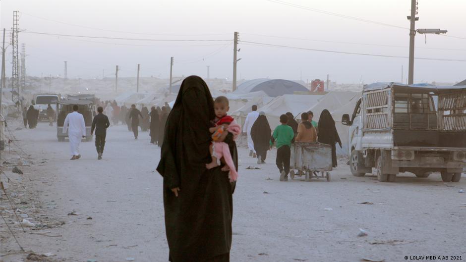 Still from "Sabaya": A woman wearing a niqab holds a child; people in the background walk through a dusty camp
