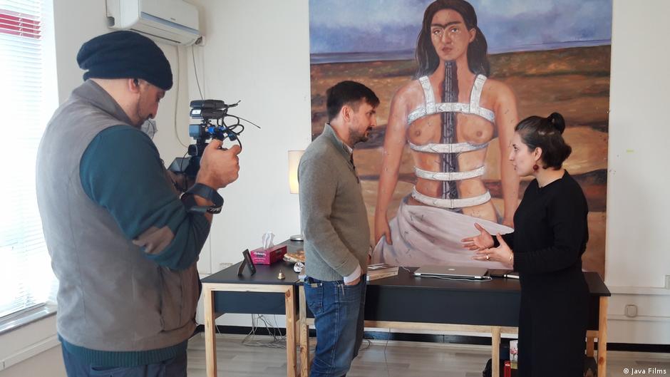 Still from "Ghosts of Afghanistan": A man talking to a woman being filmed by a third person; in the background a self-portrait of Frida Kahlo bearing her breasts