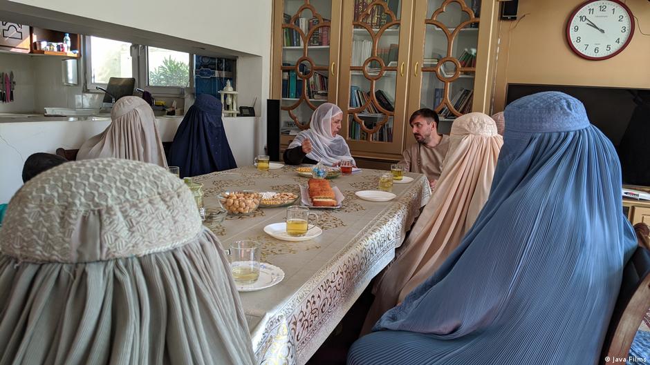 Still from "Ghosts of Afghanistan": Women in burkas sit around a table drinking tea. One woman who wears only a hijab talks to a man at the far end