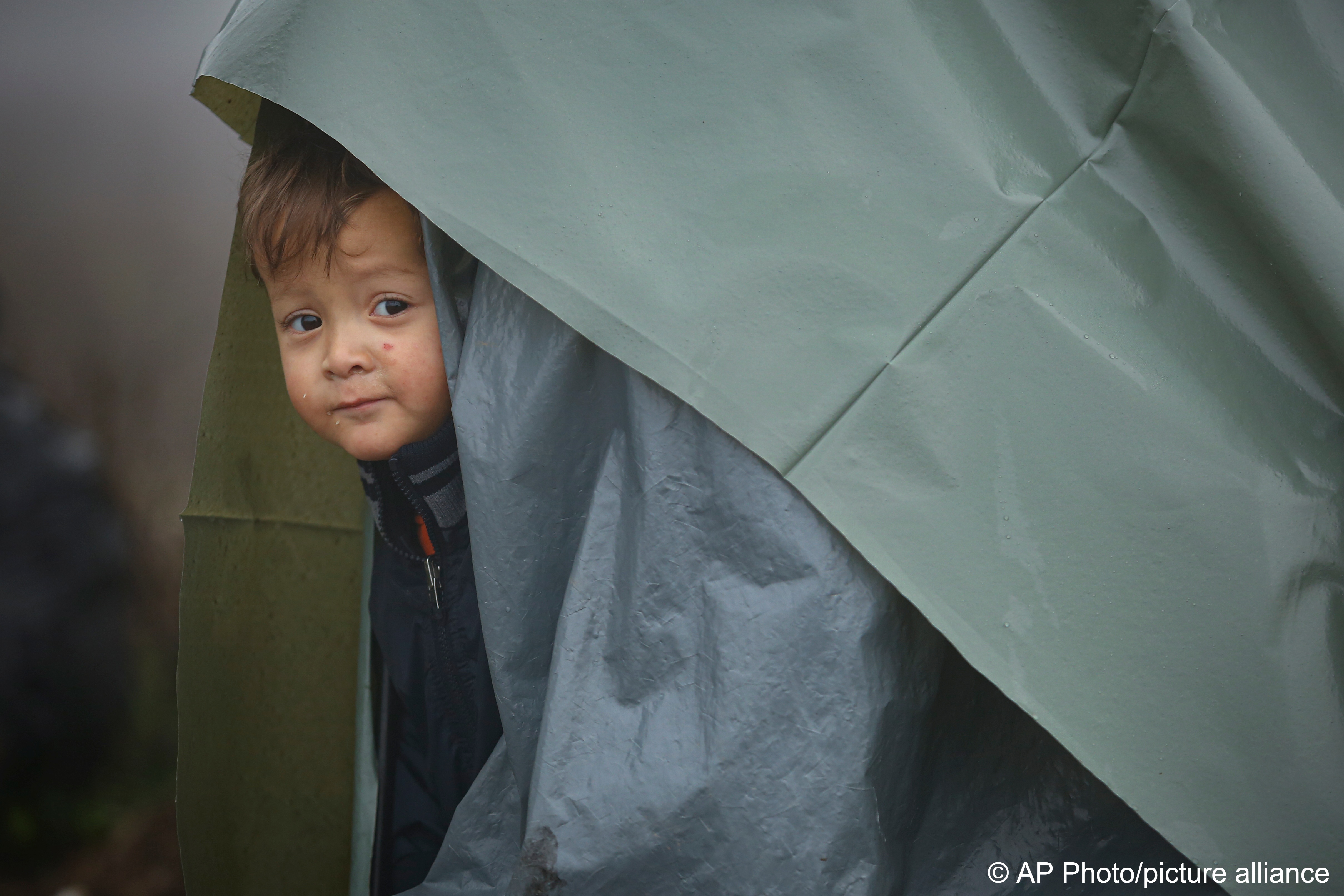 A migrant child peers from a makeshift tent at a camp housing migrants mostly from Afghanistan in Velika Kladusa, Bosnia, 12 October 2021 (photo: AP Photo)
