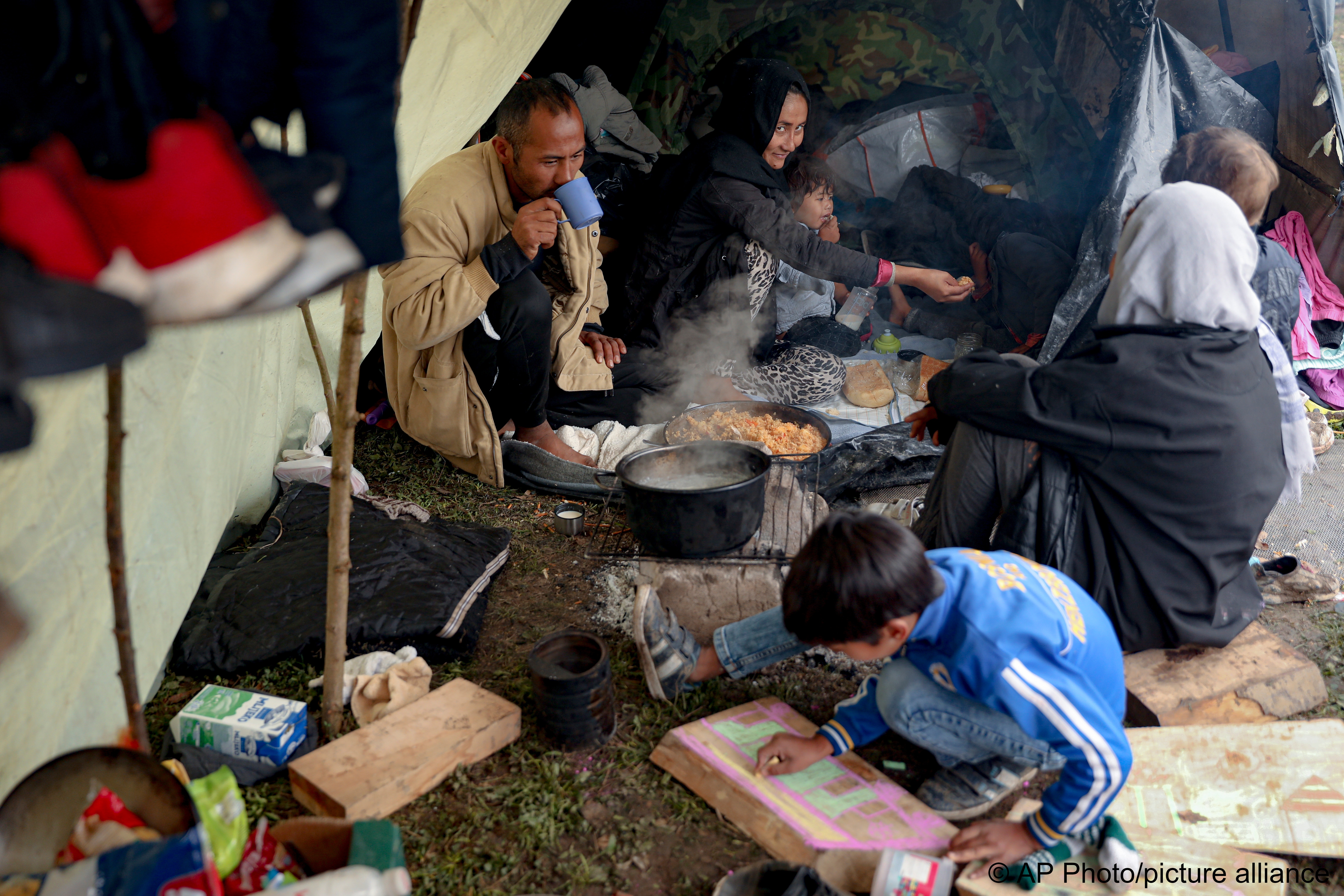 A migrant child draws on a piece of wood as people eat at a makeshift camp housing migrants mostly from Afghanistan, in Velika Kladusa, Bosnia, 12 October 2021 (photo: AP Photo)