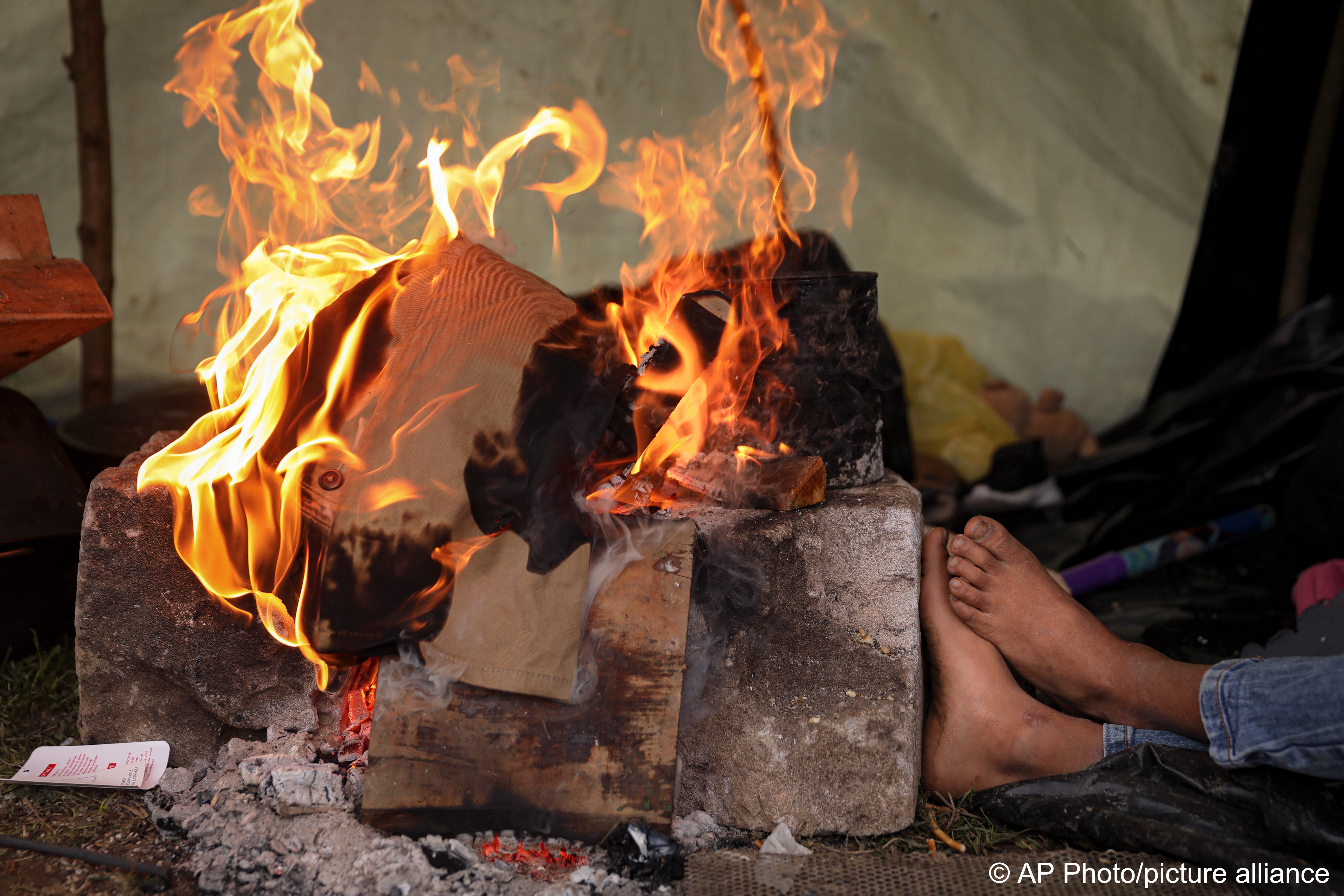 Ein Migrantenjunge wärmt sich die Füße an einem Feuer in einem Behelfslager für Migranten, die hauptsächlich aus Afghanistan stammen, in Velika Kladusa, Bosnien, 12. Oktober 2021; Foto: AP Photo