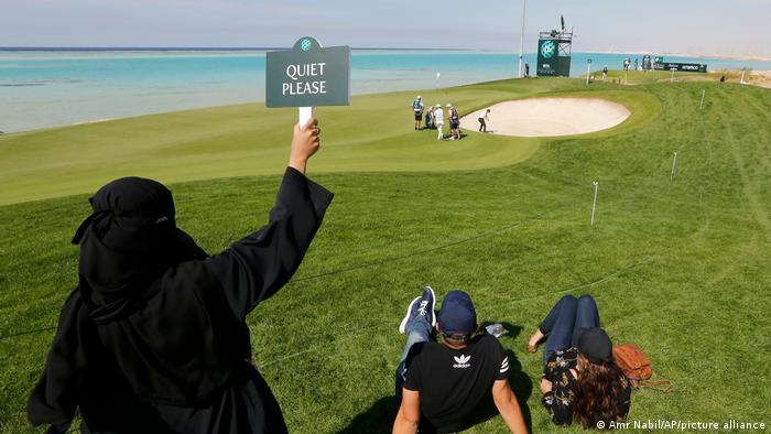 At the Saudi Golf Championship, a veiled woman on the edge of the golf course holds a sign reading 'Quiet please!'