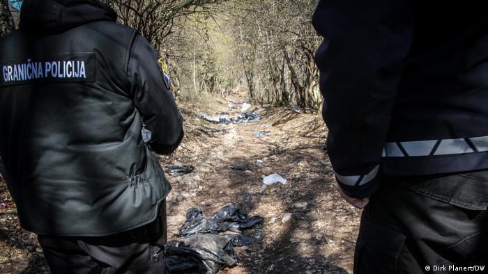 Two people are standing with their backs to the camera, on the back of the jacket of the left one is written border police in Bosnian/Croatian/Serbian, in front of the two a forest path can be seen, on which lies rubbish