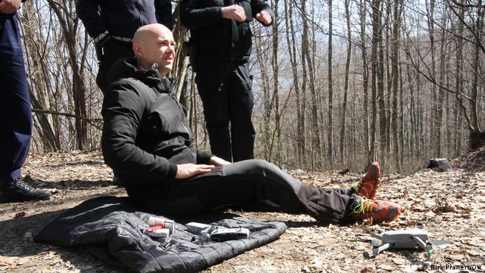 A man sits on the forest floor, next to him lies a jacket, behind him stand three other people
