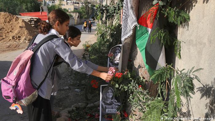 Two girls place flowers at the site where Abu Akleh was shot; pictures of the slain journalists and a Palestinian flag adorn the place