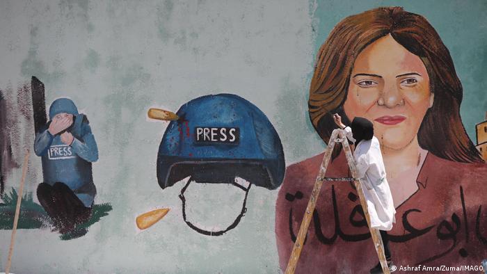 An artist paints a large mural, showing a portrait of Abu Akleh, and another depicting her with a vest and helmet, as well as a press helmet perforated by a bullet