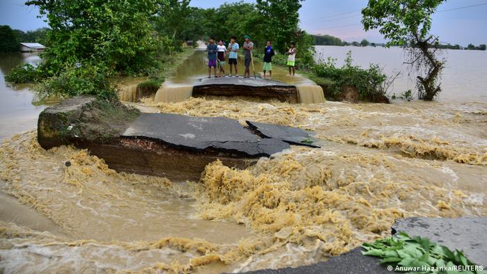 People stand behind a section of road destroyed by floodwaters in Assam, India