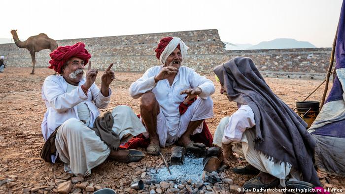 Three men with cloths on their head are sitting together in front of an extinguished fire, gesticulating and laughing. In the background is a wall, a camel and mountains