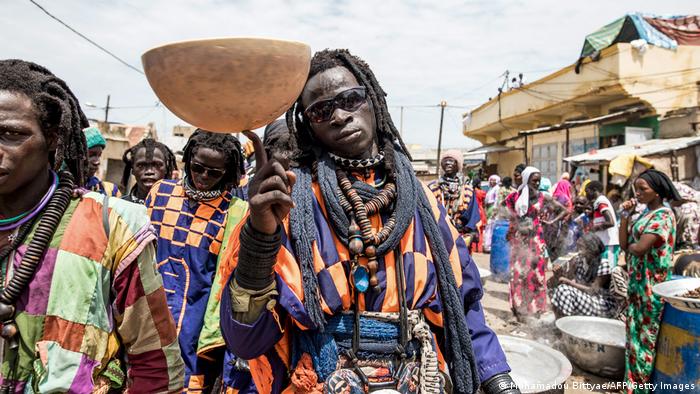 A man from the Baye Fall sect of the Mouride brotherhood wearing dark sunglasses, dreadlocks and colorful clothing, balances a bowl for collecting alms on his right index finger