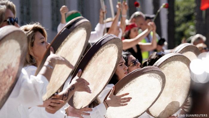 A line of women dressed in white beat large tambourine drums