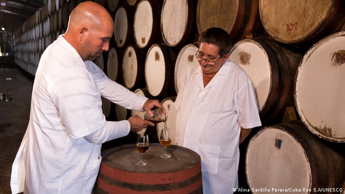 Two rum masters in white coats stand in front of barrels, one of them pours rum into a glass