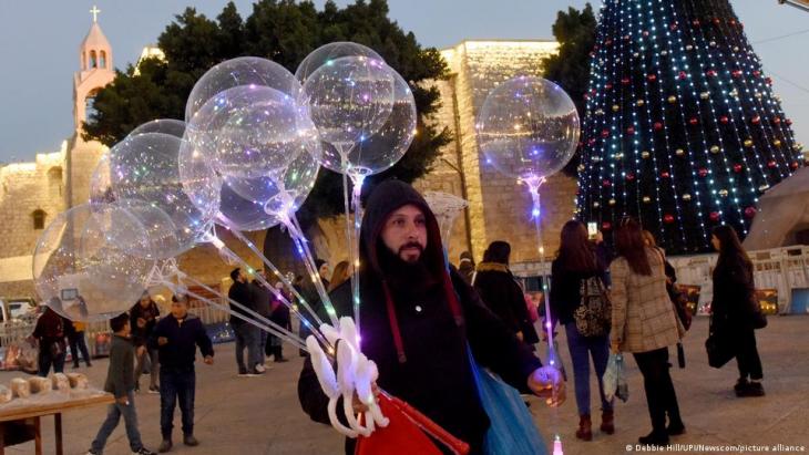 Man carrying sparkly balloons passes a Christmas tree in Bethlehem (photo: Debbie Hill/UPI/newscom/picture-alliance)