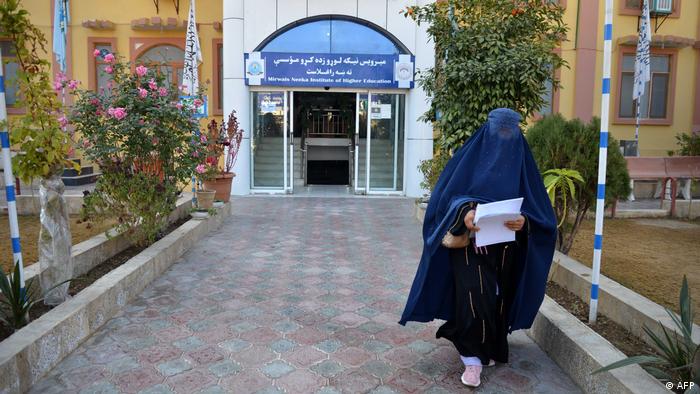 The picture shows the entrance to a university in the Afghan province of Kandahar, flanked by bushes and rose bushes. A woman clad in a dark blue burqa is walking away from the entrance, holding a stack of papers in her hand.