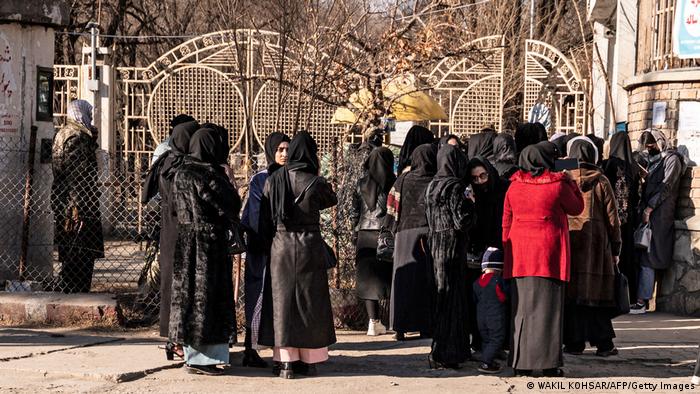 Numerous women stand in front of the closed gates of a university in Kabul, most of them dressed in black.
