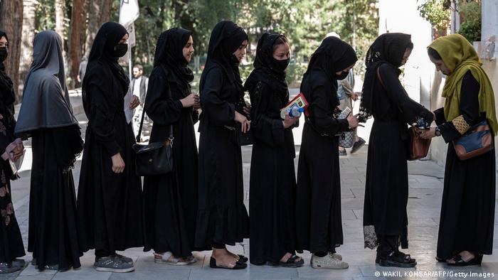 A line of young women, all in long black robes and headscarves, stand in front of Kabul University. On the right, an older woman with a green headscarf checks the bag of the young woman at the front.