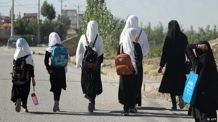 A group of girls photographed from behind are walking down a street. All are wearing dark clothes and either black or white headscarves.