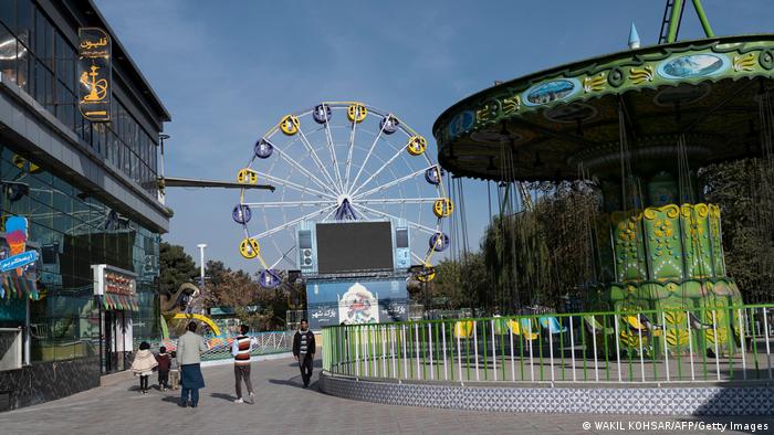 The long shot shows the largely empty City Park amusement park in Kabul. A Ferris wheel can be seen in the background and a green chain carousel on the right.