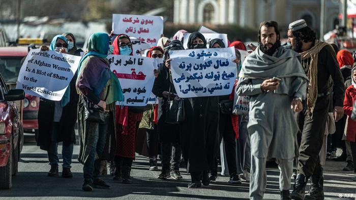 A group of women hold up banners on a busy Kabul street. On the right of the picture are two men who seem to be watching the protest suspiciously.