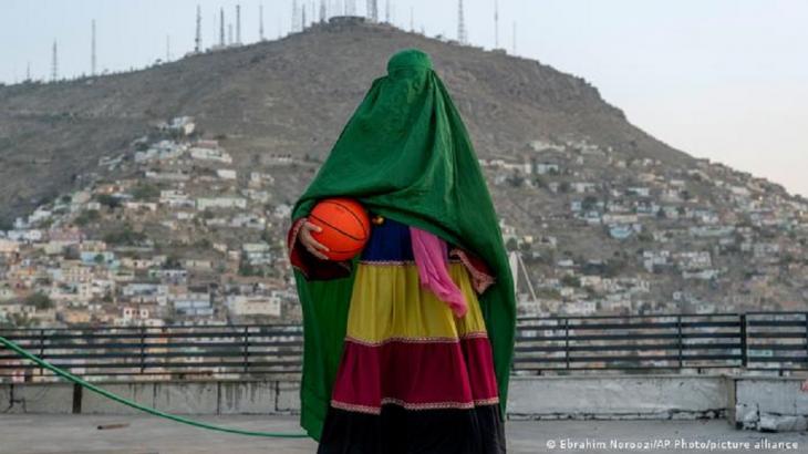 A young female basketball player stands in a burka on a public basketball court in Kabul