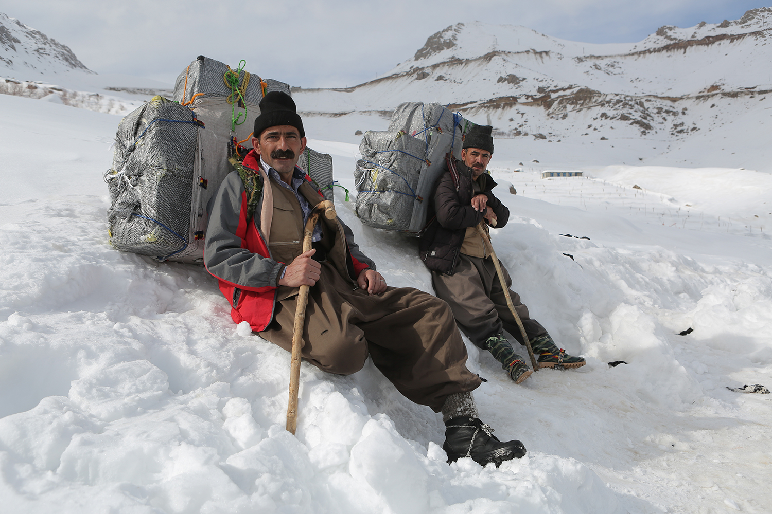 Zwei Männer mit Gepäck auf dem Rücken sitzen auf einer Schneebank; Foto: Konstantin Novakovic