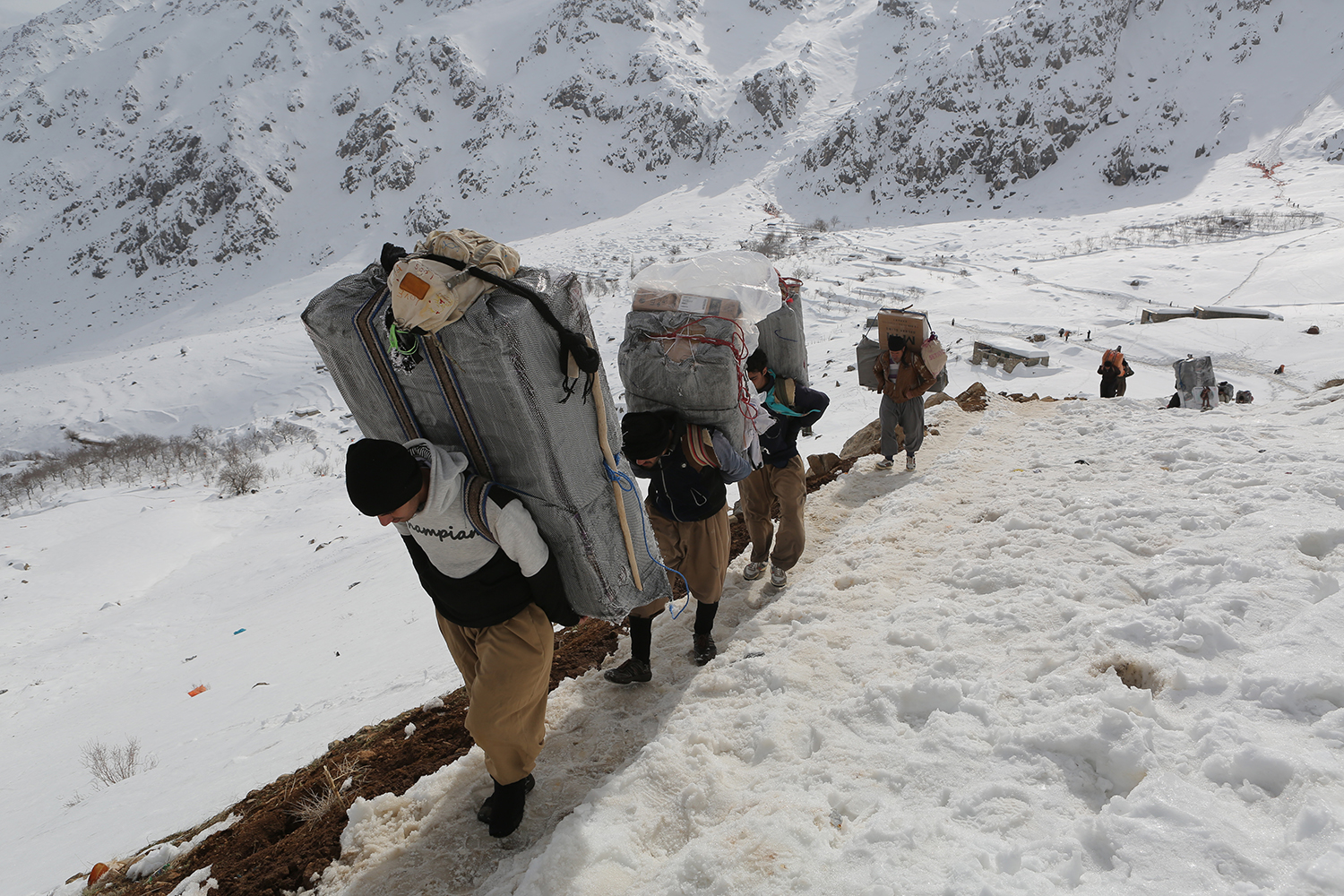 Eine Reihe von Männern geht mit riesigen Rucksäcken auf dem Rücken auf die Kamera zu, vor dem Hintergrund verschneiter Berge; Foto: Konstantin Novakovic