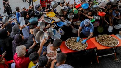 A large group of people holding empty plates, bowls and other recepticles crowd around an aid worker serving food from a large pan.