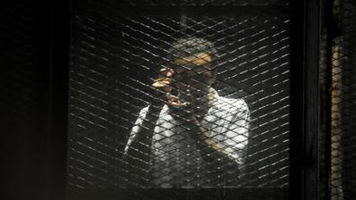 A man behind a wire panel holds his hands up to mimic taking a photograph.