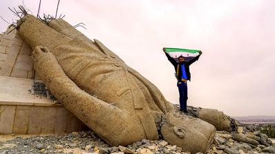 رجل يرفع علم سوريا الحرة واقفًا على رأس تمثال أسد مقلوب.