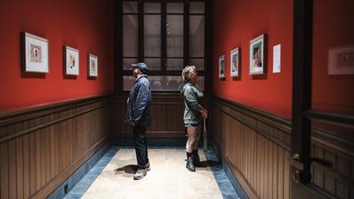 18th Istanbul Biennial (Photo: IKSV | Mete Kaan Özdilek) Two people in a corridor, facing away from eachother and looking at paintings hanging on the walls.